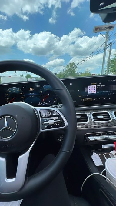 Interior view from the driver's seat of a Mercedes-Benz showing the steering wheel, dashboard with speedometer and infotainment screen, looking out at a sunny road with power lines.