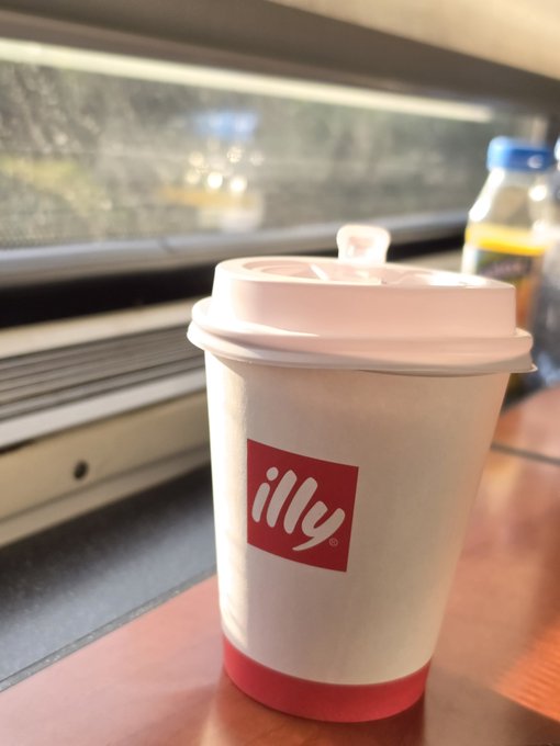 A white Illy coffee cup with its distinctive red logo sits on a wooden table beside a train window.