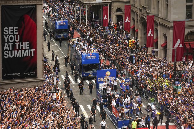 The image shows a large crowd of people gathered along a street, with two blue buses driving down the road. The buses are adorned with banners that say "Knicks in 5 New York's alive In 90 days Summit", indicating an event or celebration related to the Knicks basketball team and their journey to the summit. The scene captures the excitement and anticipation of the crowd as they watch the buses pass by, possibly waiting for the event to begin or just enjoying the sight of the buses driving down the street.

There are several people standing on both sides of the road, some closer to the buses while others stand further away. A few individuals can be seen holding handbags and backpacks, suggesting that they might have come from a nearby location such as an event or gathering. The overall atmosphere is lively and bustling with activity, reflecting the energy and enthusiasm surrounding this special occasion.