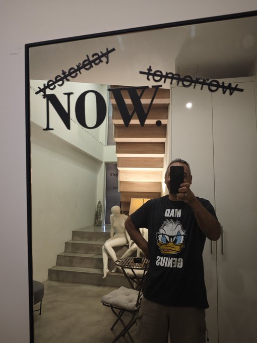 A mirror selfie of a person wearing a black 'MAD GENIUS' t-shirt with a Donald Duck graphic, standing in front of a mirror inscribed with the words 'yesterday', 'NOW.', and 'tomorrow'.