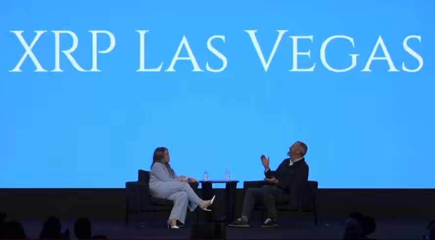 The image features a man and woman sitting across from each other at a table, engaged in conversation. They are both dressed professionally, with the man wearing a black shirt and pants while the woman has on a white dress. The background of the image is blue, which appears to be part of an event or presentation related to XRP. There are also several people visible in the background, suggesting that this could be a conference or gathering focused on cryptocurrency technology.