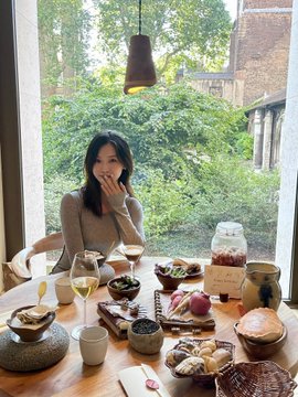 A woman laughs while sitting at a beautifully set table filled with various dishes, snacks, and drinks in front of a large window overlooking lush greenery. The scene captures a joyful birthday celebration, aligning with the post's triumphant reflection on turning 42, surviving four bear markets, and finally landing a real job—celebrating life's milestones with grace and humor.
