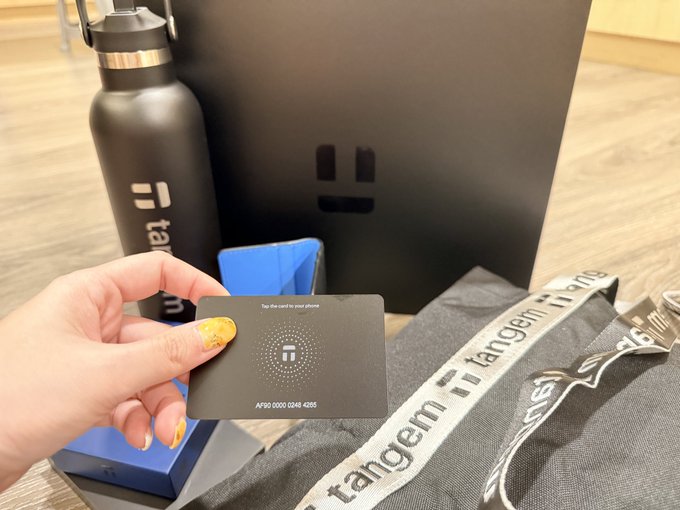 A hand holds a black 'tangem' branded card with a chip and the number 'A990 0000 0243 4205', alongside a tangem water bottle and branded packaging.