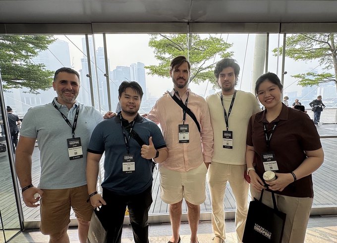 A group of five smiling people wearing 'Singapore FinTech Festival' lanyards pose for a photo indoors with large windows showing trees and a city skyline in the background.