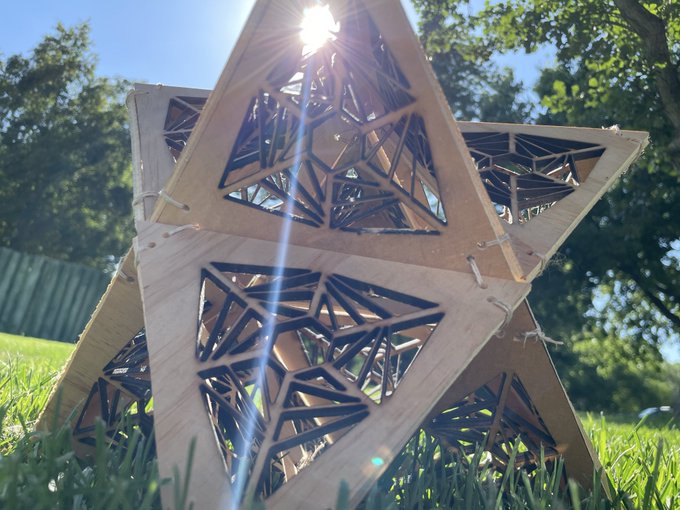 A geometric wooden star sculpture sits in grass with the sun shining through its intricate lattice patterns.