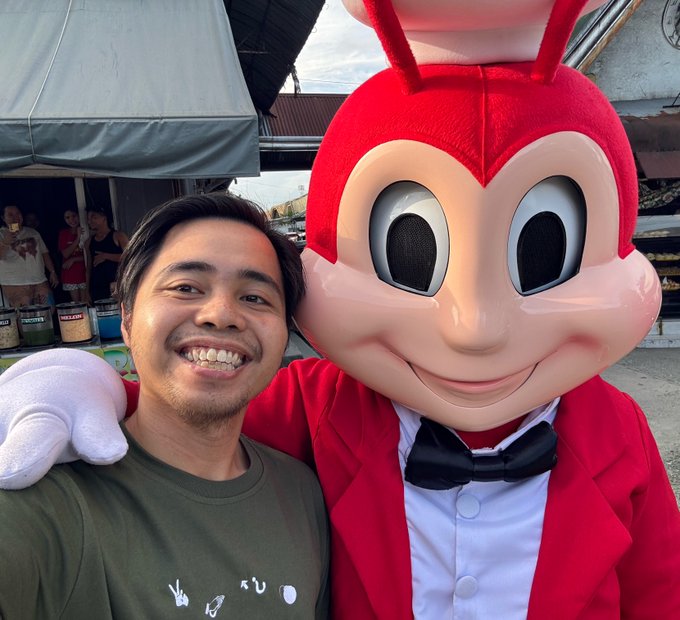 A smiling man poses for a selfie next to a person in a large red mascot costume wearing a black bowtie.