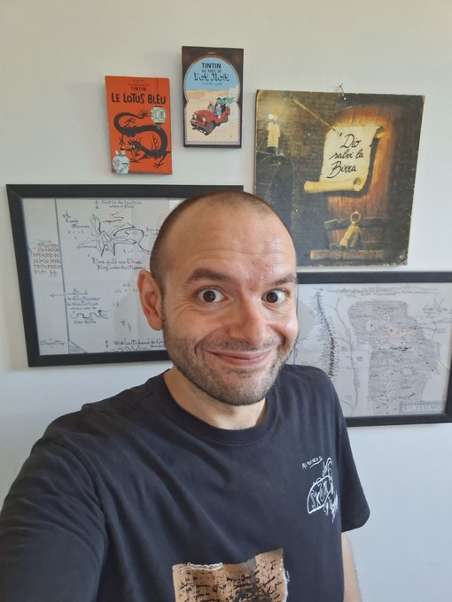 A man with a big, excited grin takes a selfie in front of a wall decorated with Tintin books ("Le Lotus Bleu," "Tintin in Tibet") and framed artwork, including one that reads "De Nacht is Brea" and another labeled "CHURFIRSTEN," capturing his enthusiastic Friday energy ahead of new video content and a "Say Something" episode with boxdotfun.