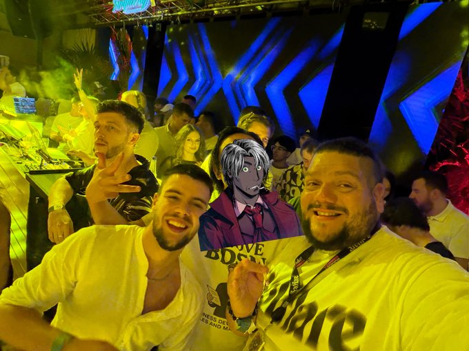 Selfie of two smiling men at a nightclub with blue and yellow lighting, one wearing a white shirt and the other wearing a shirt with 'Affiliate' visible, with a cartoon character edited in between them.
