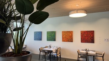 A restaurant or cafe interior with a large potted plant in the foreground, a round pendant light, and colorful abstract paintings on a white wall behind small dining tables.