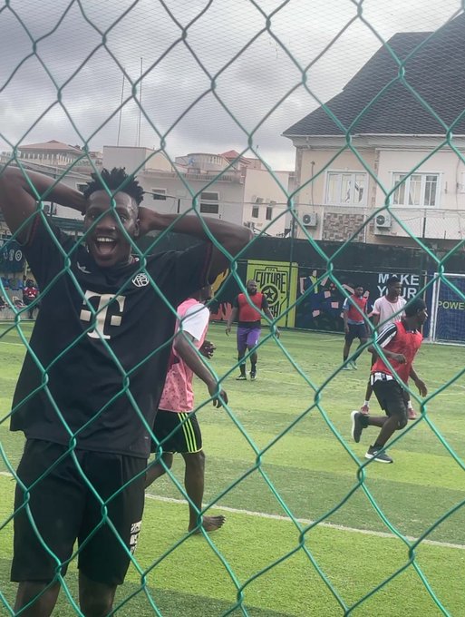 A young man with his hands behind his head smiles broadly through a green chain-link fence while watching a soccer match, with a banner reading "CT FUTURE" visible in the background—matching the "CT" referenced in the post about him showing off his ball skills.