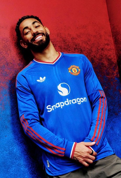A man smiles with closed eyes while leaning against a red and blue backdrop, wearing a blue Manchester United jersey featuring the Snapdragon sponsor logo and Adidas trefoil. The relaxed, joyful pose and vibrant colors align with the "AURA" theme, capturing a confident, positive energy.
