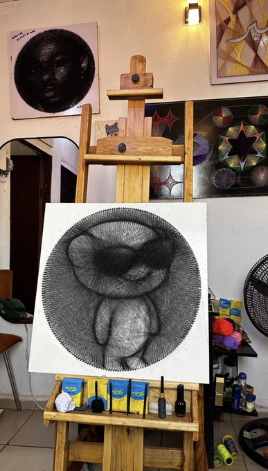 A black and white string art piece depicting a face in profile is displayed on a wooden easel, with tubes of paint on the shelf below and other artworks visible on the walls.
