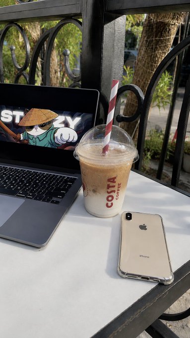 A Costa Coffee iced drink with a red and white striped straw sits on a table next to a laptop and a gold iPhone at an outdoor café.