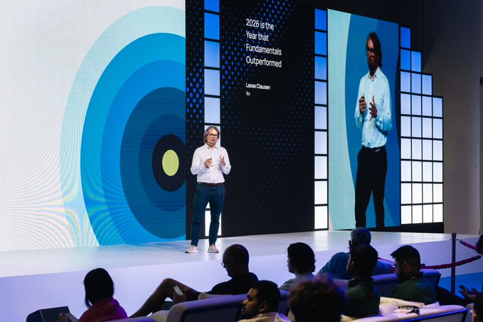 A man stands on a stage giving a presentation in front of a large screen displaying a quote: '2026 is the Year that Fundamentals Outperformed' by Lasse Clausen, while an audience watches from below.