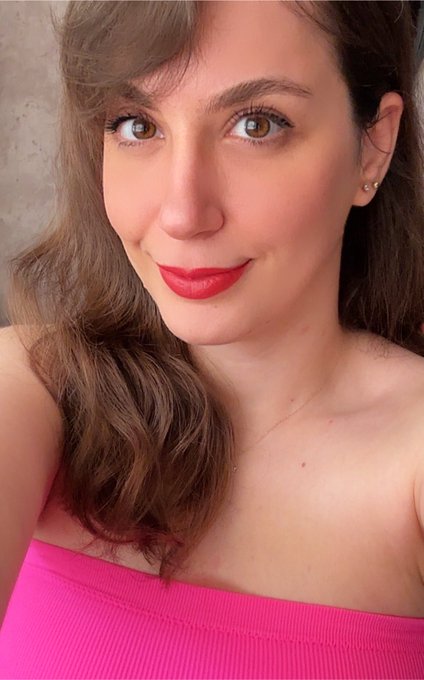 A close-up selfie of a woman with brown hair wearing red lipstick and a pink strapless top, with small stud earrings visible.