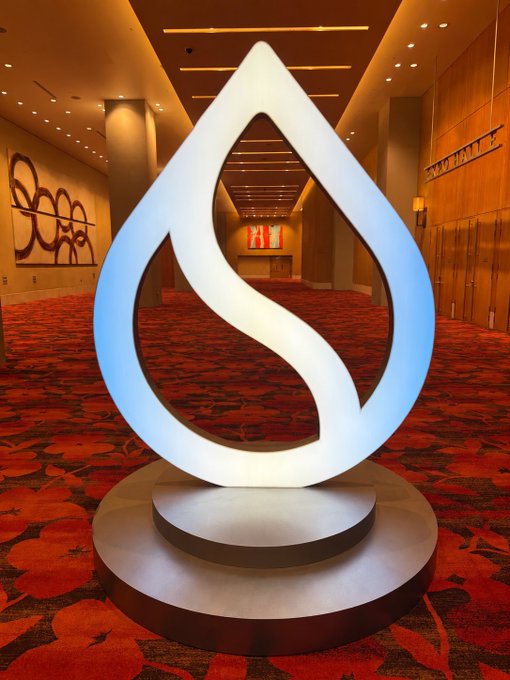 A luminous teardrop-shaped sculpture with an internal S-curve design stands on a pedestal in a carpeted hotel hallway with floral patterned rugs.