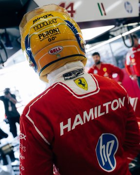 A Ferrari driver wearing helmet text "DXC," "Thermo Treat," "PERONI 0.0%," and "BELL" stands in the pit lane with "HAMILTON" printed on the back of his red race suit, linking to the post's theme of following Scuderia Ferrari's pursuit of qualifying performance.