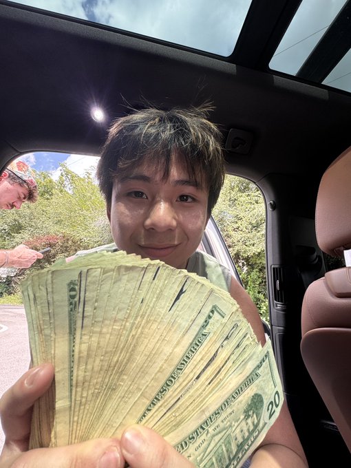 A young man sits inside a car holding up a fan of US $20 bills to the camera, smiling proudly. The image illustrates the post's message of giving back, as the cash represents the charitable act of blessing others "one $ANSEM at a time."