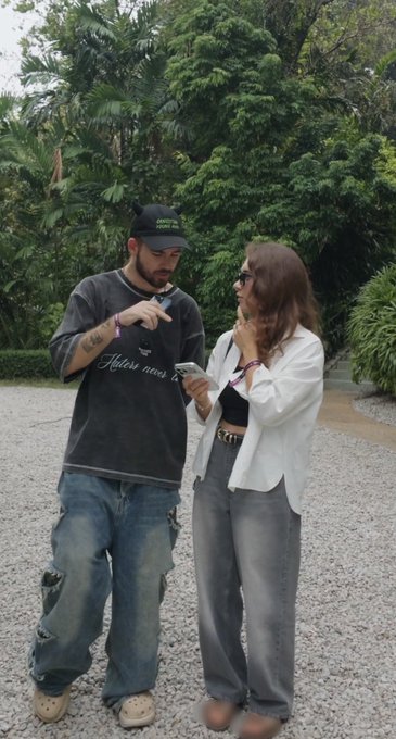 A man in a black t-shirt and jeans points at a smartphone held by a woman in a white top and gray jeans standing on a gravel path with greenery in the background.
