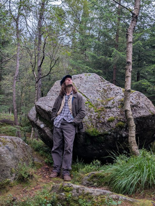 A man wearing a dark cap, plaid shirt, and jacket stands in a forested area, looking up at the sky next to a large mossy boulder.