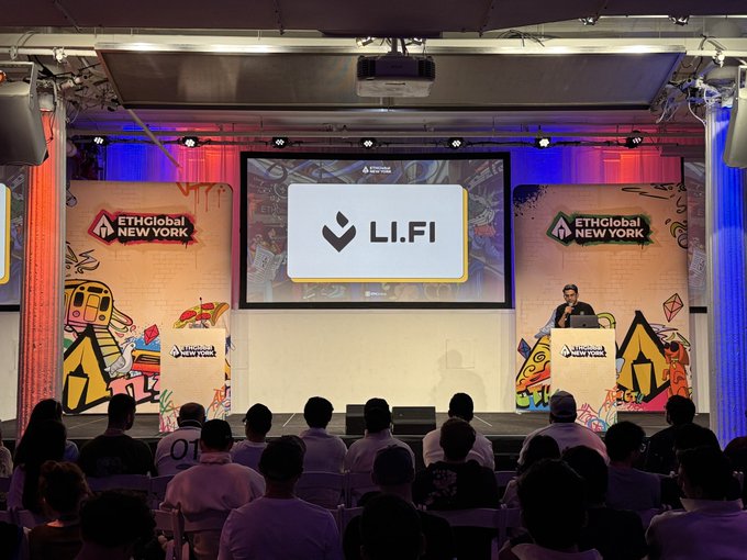 The image shows a large room with a stage set up for an event, featuring a projector screen displaying the logo "L.I.F.I". A man is standing behind a podium at the front of the room, likely presenting or speaking about the company's mission and goals. The audience consists of several people seated in chairs around the stage, attentively listening to the presentation.