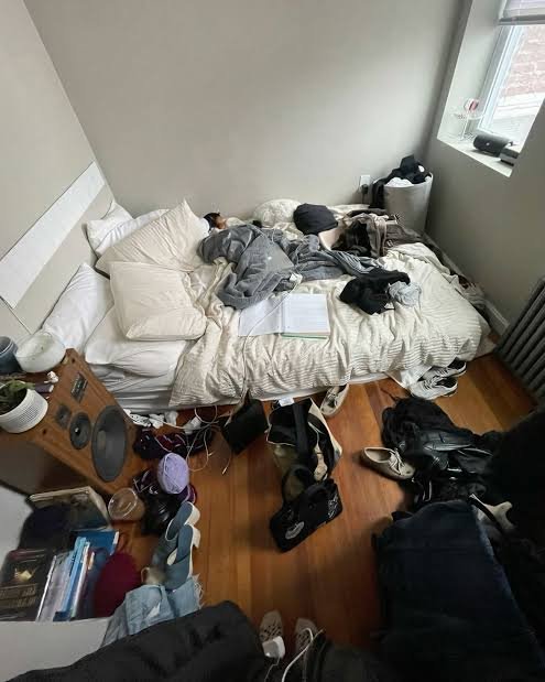 A photograph of a very messy bedroom with clothes, bags, and various items scattered across a rumpled bed and the wooden floor.