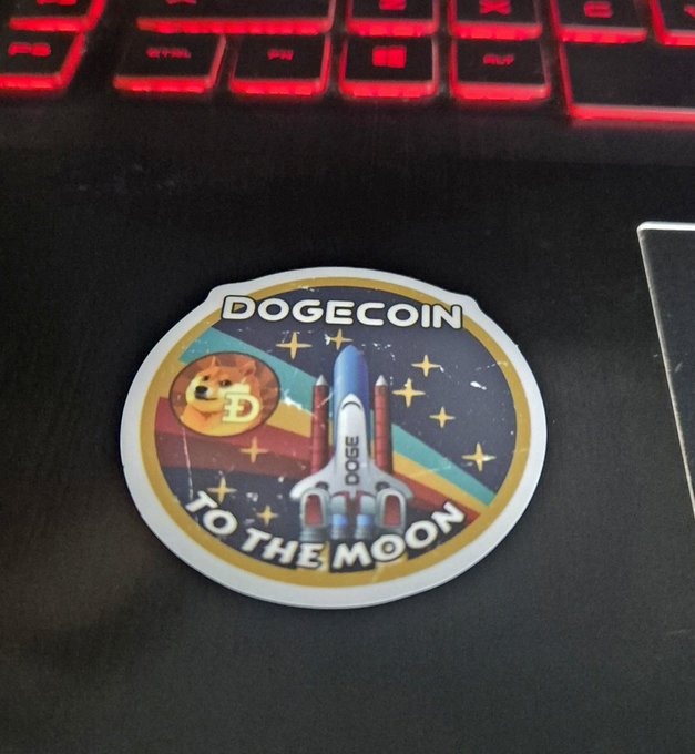 A 'DOGECOIN TO THE MOON' sticker with a rocket and Shiba Inu dog illustration sits on a black surface near a red-backlit keyboard.