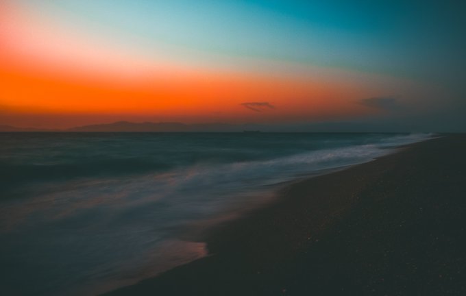 A serene beach at sunset features a gradient sky of orange and deep blue over calm waters, with distant mountains silhouetted on the horizon and smooth waves lapping onto a pebbled shore. The image embodies "Aqua Aeterna," a timeless and ethereal seascape that evokes the peaceful stillness of dawn greeting the morning.
