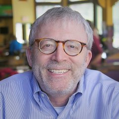 A smiling older man with gray hair, a beard, and tortoiseshell glasses wearing a blue striped shirt appears to be Peter Brandt, the trader referenced in the post predicting Ethereum's long-term price target of $8,600 after clearing the $5,000 resistance level.