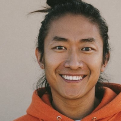 A headshot photo of a smiling young man with dark hair pulled back, wearing an orange hoodie.