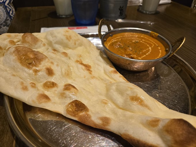 A large piece of naan bread served on a metal tray alongside a copper bowl of orange curry, likely butter chicken or tikka masala, on a wooden restaurant table.