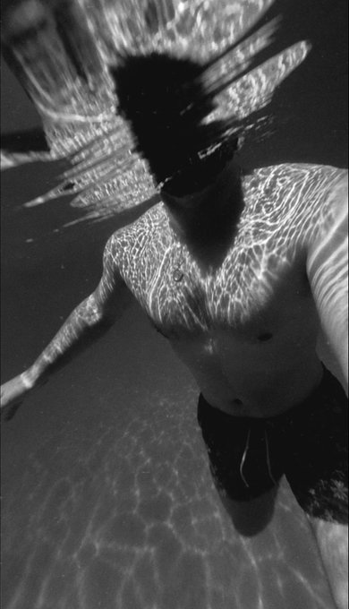 A black-and-white underwater photograph of a man in swim trunks, with caustic light patterns dancing across his torso and face obscured by ripples.