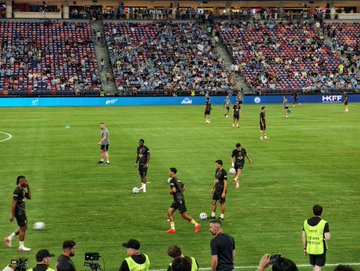 A view of a soccer field during a match, showing players in dark uniforms warming up on the green pitch while stadium spectators and 'HKFA' signage are visible in the background.