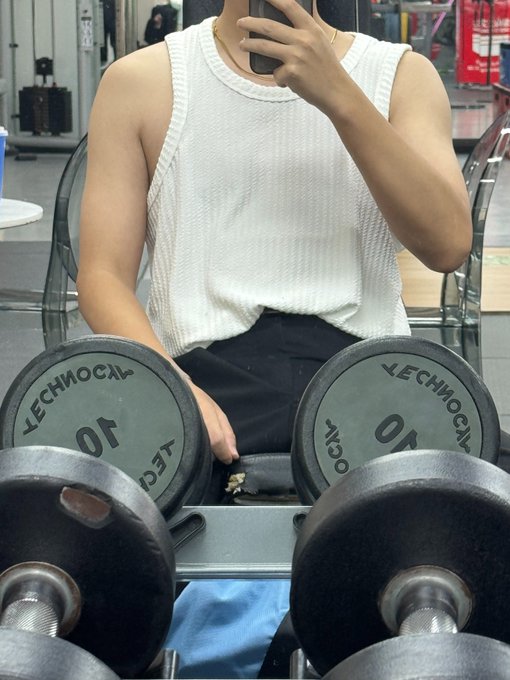 A muscular person in a white tank top sits on a gym bench taking a mirror selfie, with two "Technogym 10" dumbbells prominently placed in the foreground, while the accompanying post humorously compares their gains to crypto trading success, referencing an "11M take-profit price" and tagging a Solana contract address.