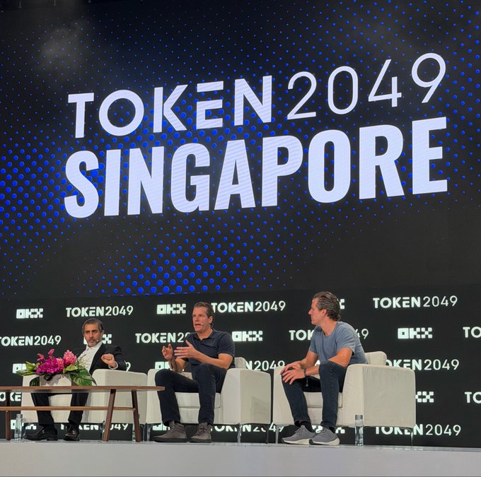 Three men sit on white armchairs on stage at TOKEN2049 Singapore, with a large screen behind them displaying the event name, as they engage in a panel discussion. The image visually supports the post's highlight of Zcash ($ZEC) co-founders Tyler and Cameron advocating for privacy at the crypto conference.