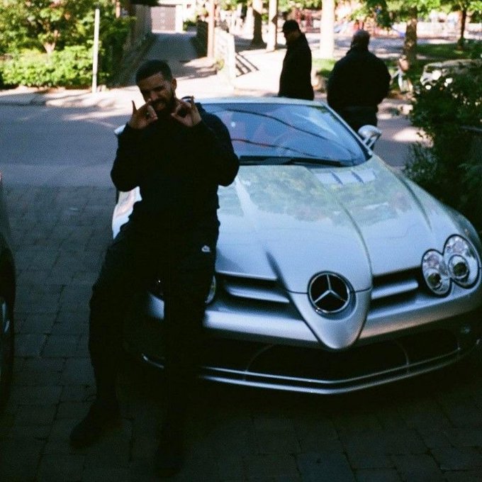 Drake poses with both hands making 'OK' gestures in front of a silver Mercedes-Benz SLR McLaren convertible on a brick pathway, with two security guards standing in the background.
