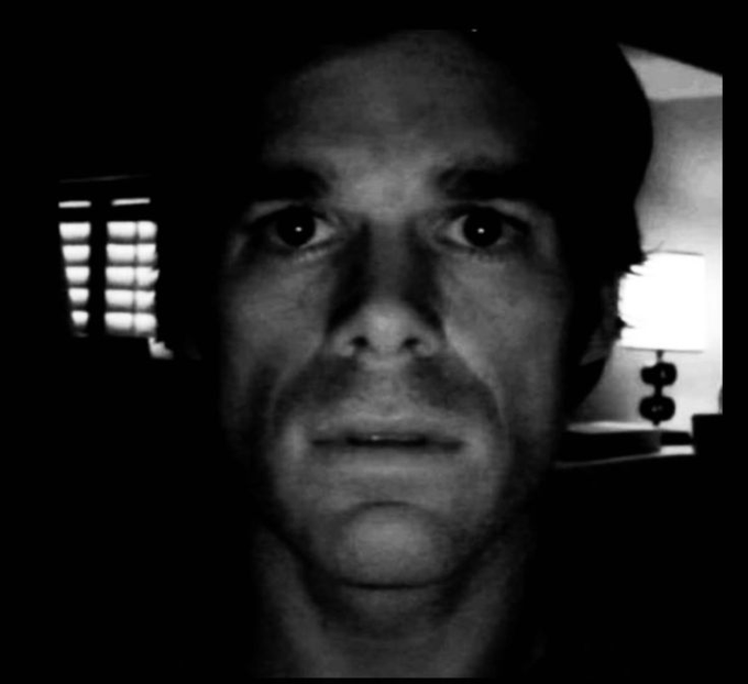 A stark black and white close-up photo captures a man with an intense expression looking directly at the camera, with window blinds and a lamp visible in the dimly lit background.