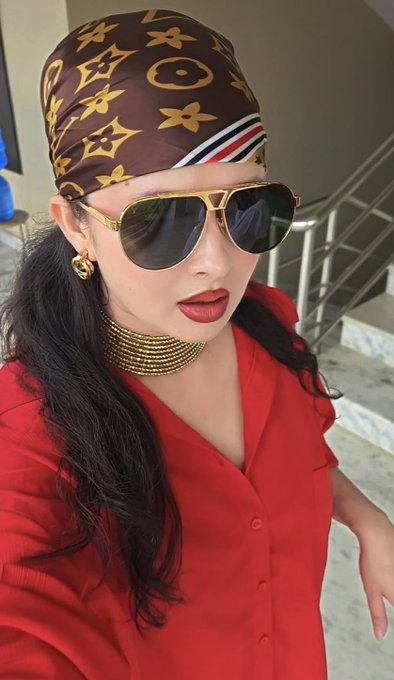 A selfie of a woman wearing a red top, a gold choker, sunglasses, and a brown patterned headscarf.