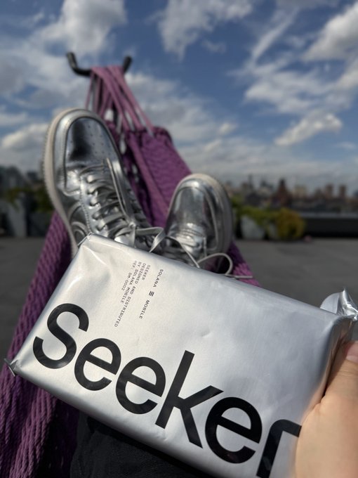 Casual outdoor photo showing silver sneakers resting on a person in a purple hammock, with a silver Seeker package held in the foreground against a city skyline.