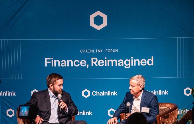 Two men in suits seated on stage having a discussion at the Chainlink Forum, with a large blue backdrop reading 'CHAINLINK FORUM Finance, Reimagined'.