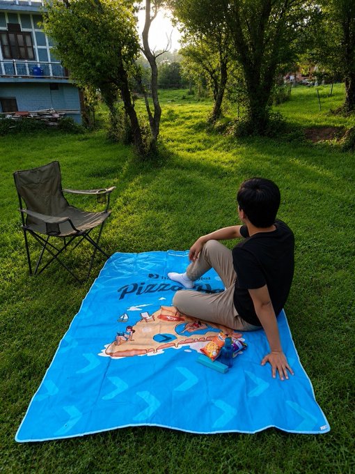A young person relaxes on a blue "Pizza" beach mat in a grassy area, with a folding chair nearby, embodying the freedom and mobility that the Bitget Wallet message promotes. The serene outdoor setting reinforces the idea of taking your crypto wallet on-the-go wherever the weekend leads.