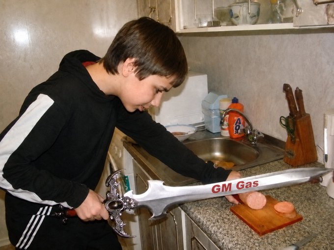 A young boy in a black and white hoodie stands in a kitchen, dramatically wielding a large decorative sword that reads 'GM Gas' as he prepares to cut a sausage on a wooden cutting board.