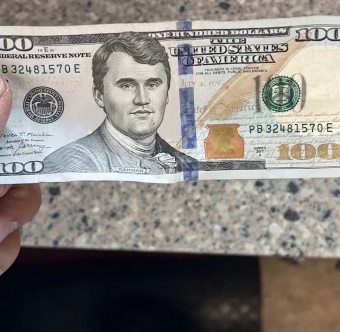 A close-up of a US $100 bill featuring a stylized portrait labeled 'RAHMAN' instead of the typical Benjamin Franklin, held by someone's fingers against a speckled granite-like background.