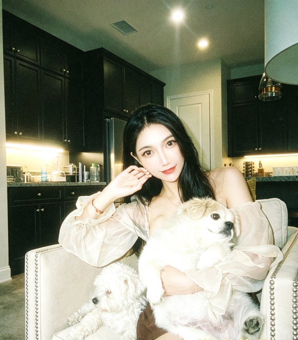 A selfie-style photo of a young woman in a sheer light-colored top sitting in a cream armchair with two small fluffy white dogs, in a dimly lit kitchen with dark cabinets.