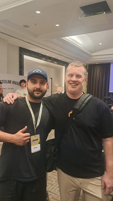 The image shows two men standing next to each other with their arms around one another's shoulders. They are both smiling for a photo, suggesting that they have had an enjoyable time at the Stellar hackathon or party they attended recently. The event was described as incredible and filled with talented people from the Stellar team who provided feedback on various projects. One of the men in the image is wearing a name tag, indicating their participation in the event.