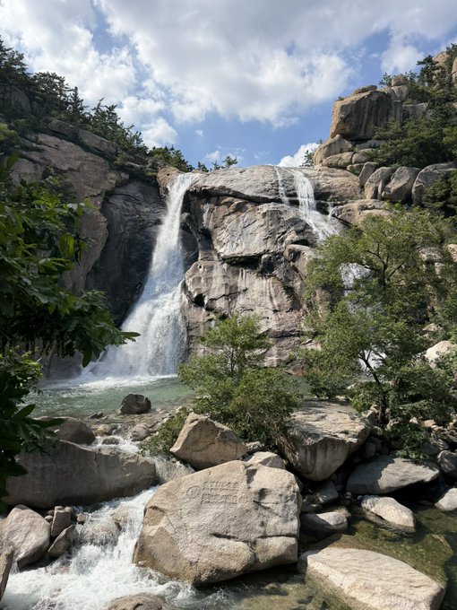 A powerful waterfall cascades down rocky cliffs into a turbulent pool below, with a large boulder in the foreground bearing faint engraved text that reads "BISHOP" and other weathered markings. The image's rushing water and rugged landscape mirror the post's reflection on trading—natural forces surging forward relentlessly, much like how concentrated bets can crash over a trader who chooses to hold motionless through the fall.