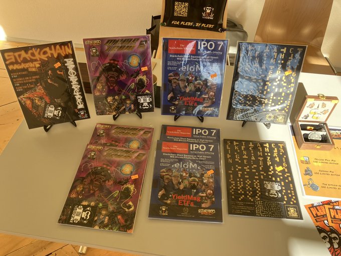 A tabletop display of Yu-Gi-Oh! merchandise includes visible titles “STACK CHAIN,” “The Violets Magician,” “Gladiator's Assault,” “IPO 7,” and “Yu-Gi-Oh! TRADING CARD GAME,” along with a QR-code sign reading “FOR PLEASE, BE PLEASE.”