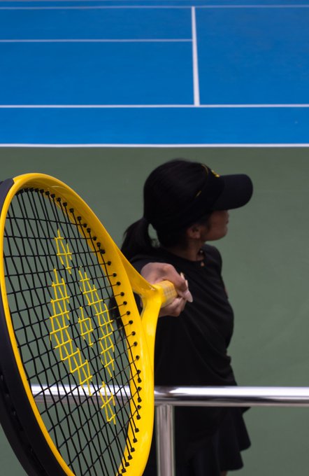 A female tennis player wearing a black cap and outfit stands on the court holding a large yellow tennis racket with "YONEX" branding visible on the strings. The image appears to be a stylized tennis photograph, making a simple caption unnecessary as the post indicates.
