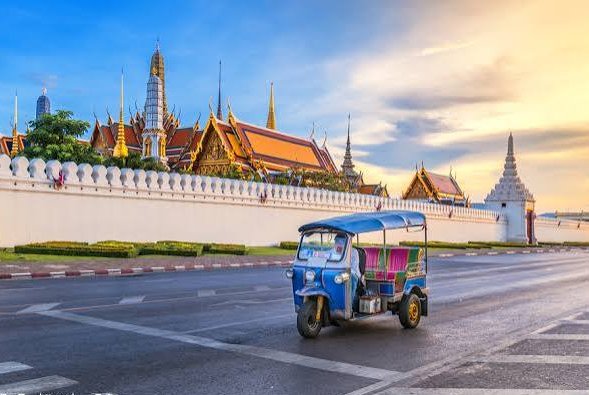 A blue tuk-tuk parked on a road in front of an ornate Thai temple complex with golden spires under a partly cloudy sky.