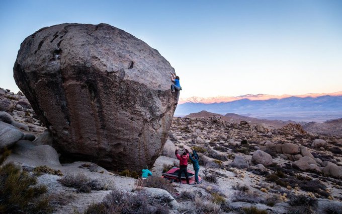Outdoor photograph of people bouldering and spotting a climber on a massive boulder in a desert landscape with mountains in the background.